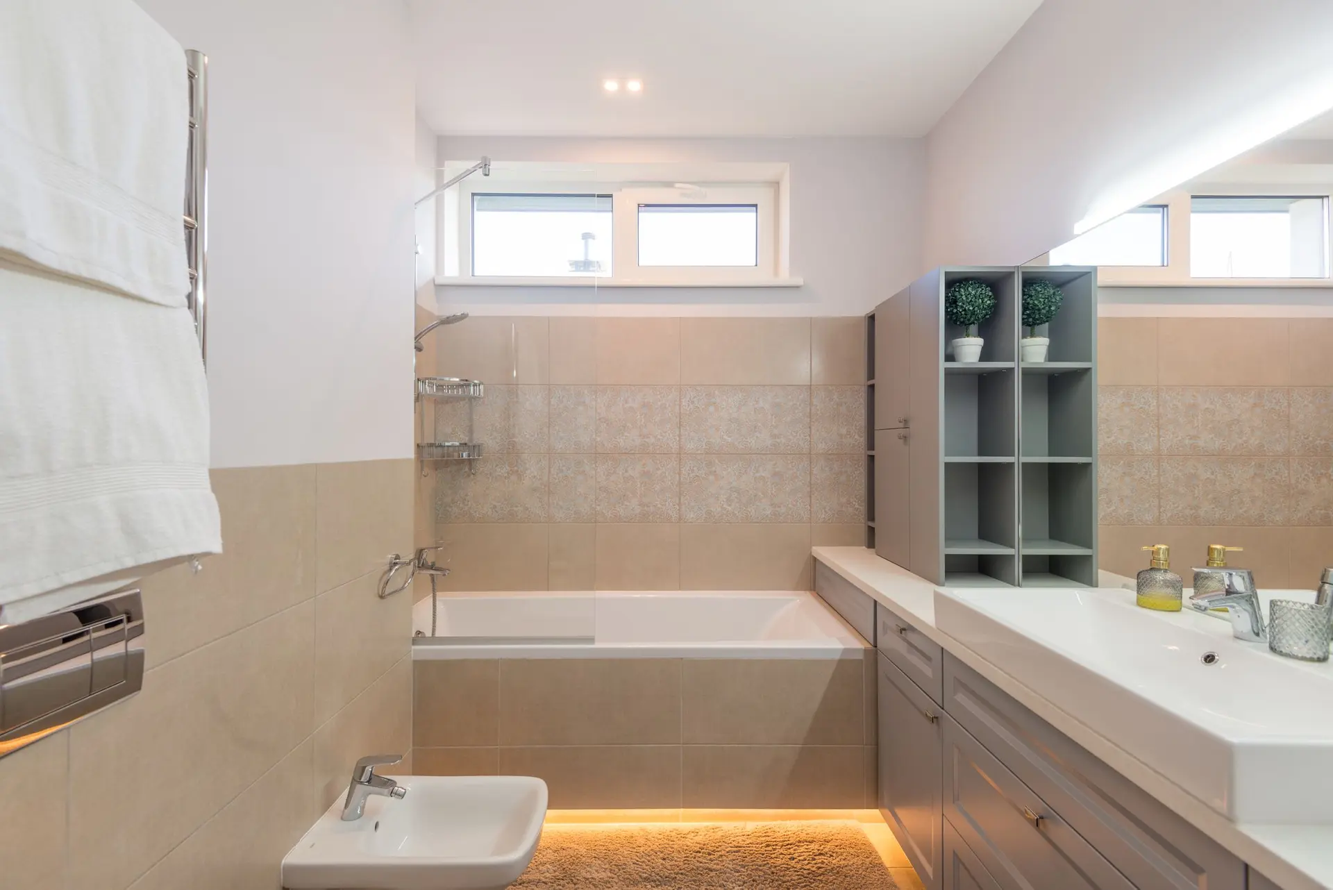 Baño en tonos arena con bañera empotrada, lavabo doble y luz indirecta bajo el faldón