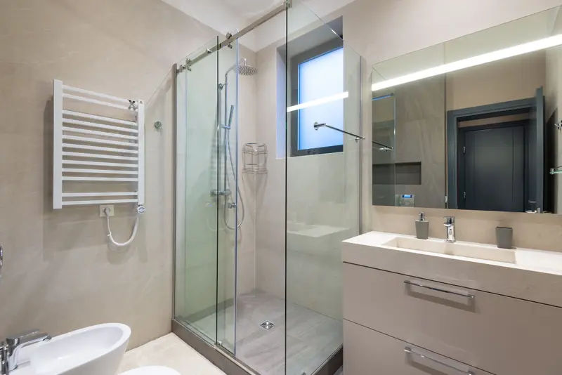 Ducha con mampara de cristal corredera sobre plato gris con desagüe central, alicatado beige en gran formato, ventana pequeña de cristal translúcido, toallero blanco en la pared y lavabo con mueble a la derecha