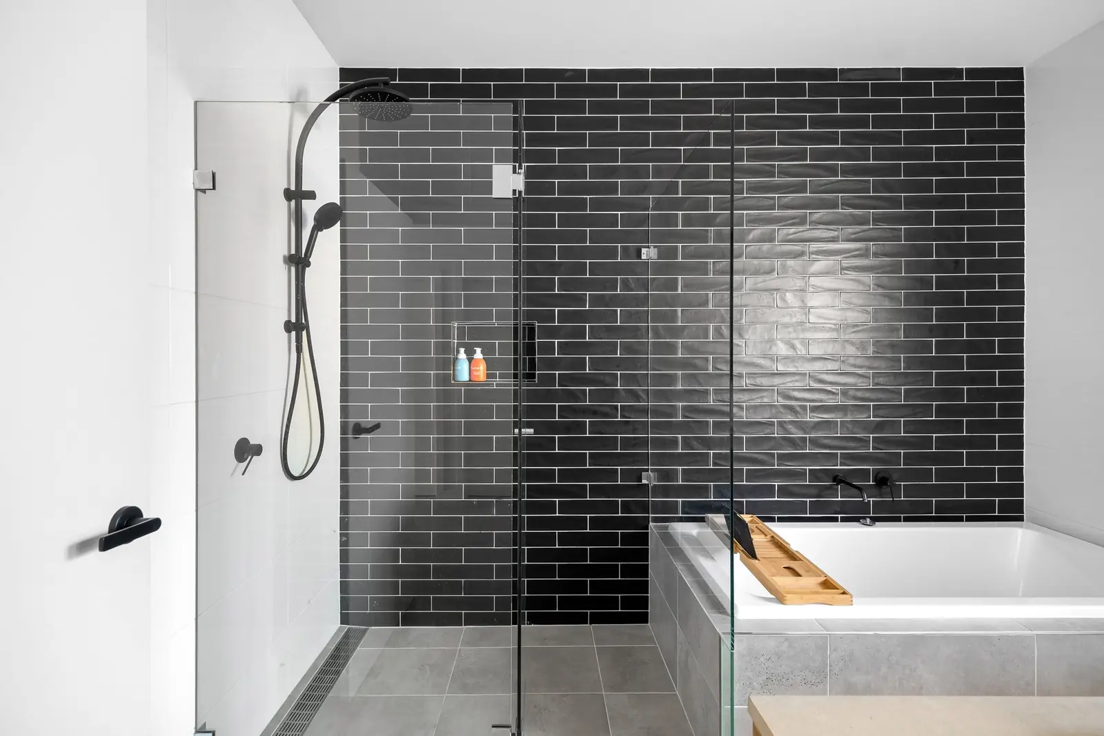 Ducha a ras de suelo con sumidero lineal junto a la pared, mampara de cristal fijo sin perfil, grifería negra con rociador y teleducha, hornacina en la pared de azulejo negro alargado y una bañera exenta al fondo a la derecha