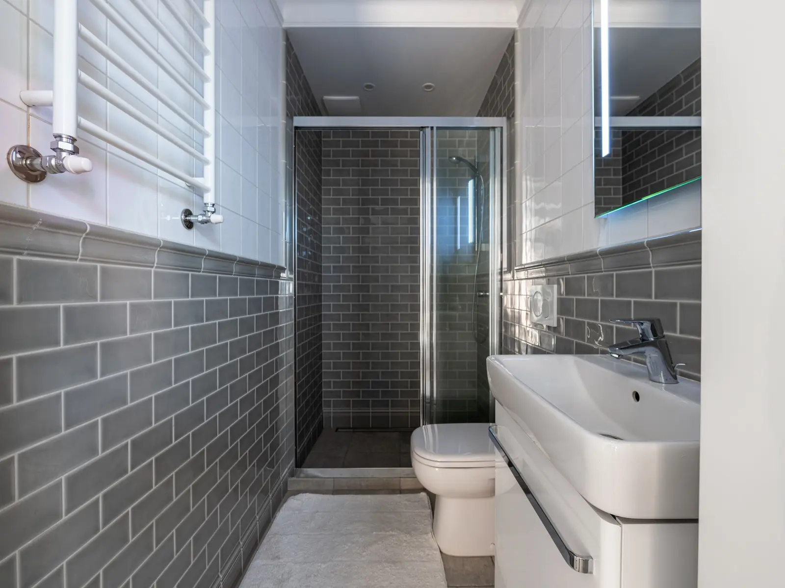 Baño estrecho con azulejo tipo metro gris en la parte baja de las paredes, ducha con mampara corredera al fondo, inodoro, lavabo con mueble blanco y toallero de agua en la pared