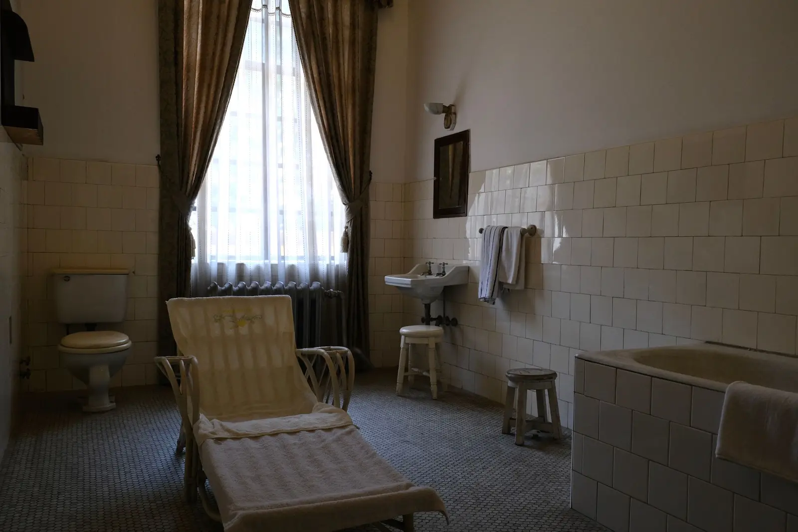 Baño antiguo con azulejo blanco cuadrado hasta media altura, bañera alicatada en primer plano, lavabo de pie, inodoro con cisterna baja junto a una ventana alta con cortinas y suelo de mosaico pequeño
