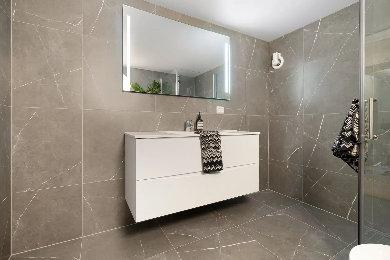 Baño revestido de suelo a techo con porcelánico gris veteado en gran formato, mueble de lavabo blanco suspendido, espejo con luz integrada y mampara de cristal a la derecha