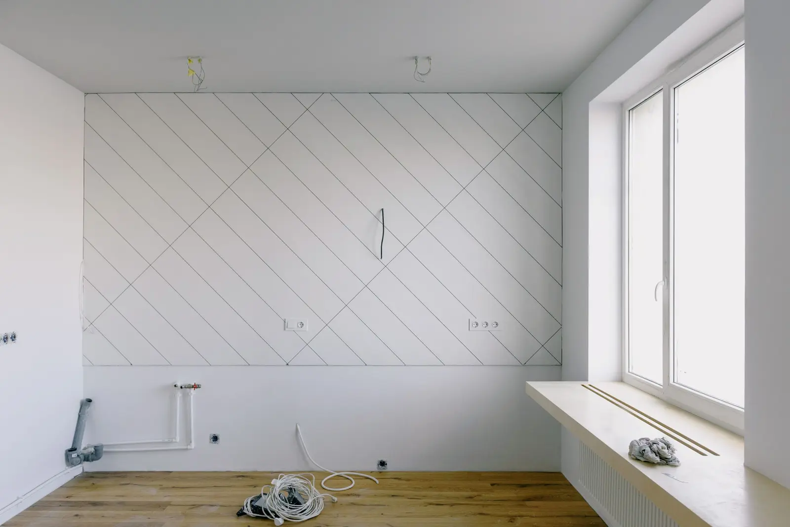 Pared de cocina alicatada en diagonal con piezas blancas, cables asomando para la campana, dos cajas de enchufe, tomas de agua y desagüe gris junto al suelo de madera, antes de montar los muebles