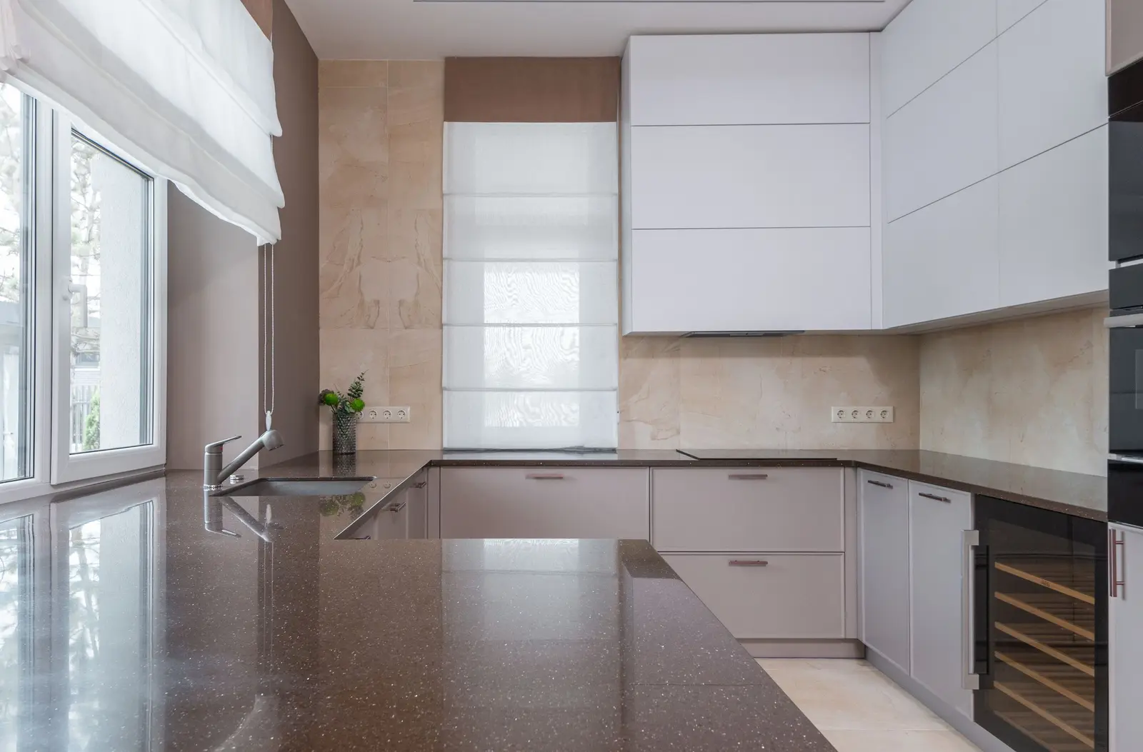 Cocina en U con muebles blancos sin tiradores, encimera oscura de piedra con motas brillantes, fregadero bajo la ventana y frente revestido de piezas color crema