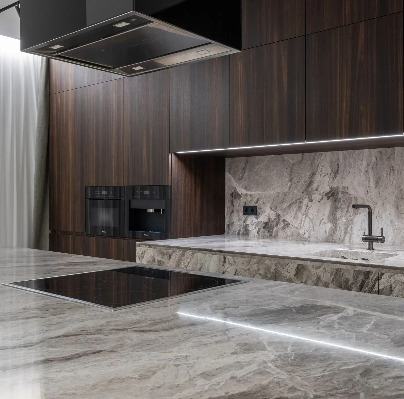 Isla de cocina con encimera de piedra gris veteada, placa de inducción, campana negra suspendida del techo, muebles de madera oscura y dos hornos en columna