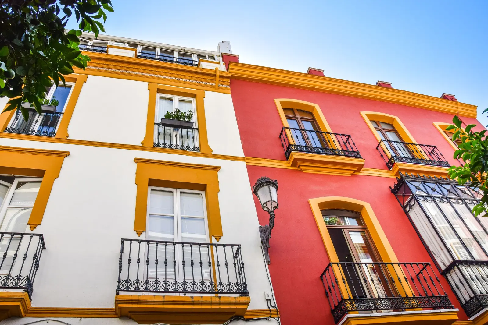 Fachadas de dos edificios de viviendas vistas desde abajo, una blanca y otra roja, con recercados amarillos en las ventanas, balcones de forja y una farola en la esquina