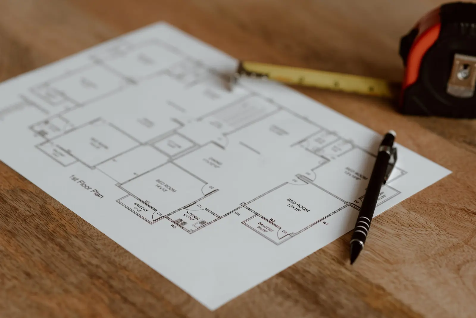 Plano en papel de la planta de una vivienda sobre una mesa de madera, con un bolígrafo negro encima y un metro de cinta amarillo y naranja al fondo
