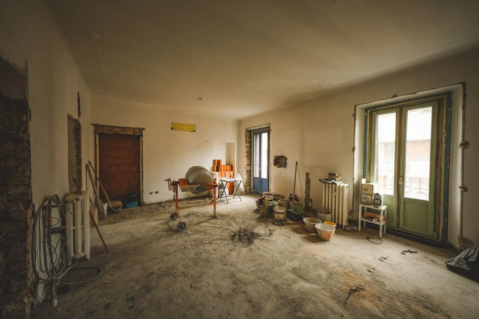 Salón de un piso antiguo en plena obra, con el suelo picado cubierto de polvo, una hormigonera, cubos, ladrillos apilados, un radiador de hierro y puertas balconeras de madera