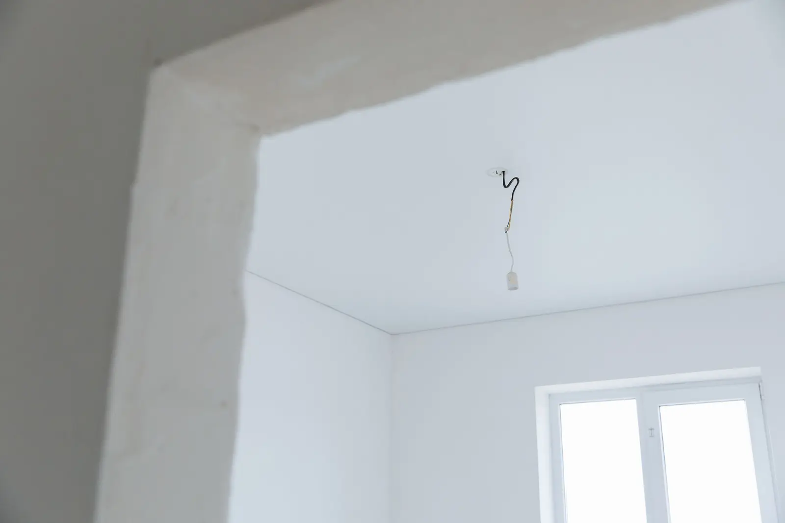 Hueco de puerta con el yeso sin rematar y, al fondo, una habitación de paredes blancas con un cable eléctrico colgando del techo para la futura lámpara, junto a una ventana