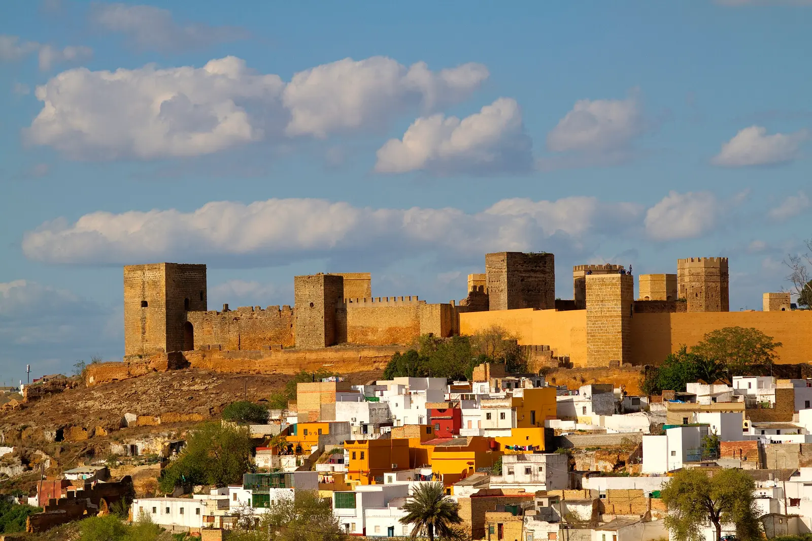 Castillo almohade de Alcalá de Guadaíra sobre el cerro, con sus torres de piedra dorada, y a sus pies el barrio de casas blancas, amarillas y ocres escalonadas en la ladera