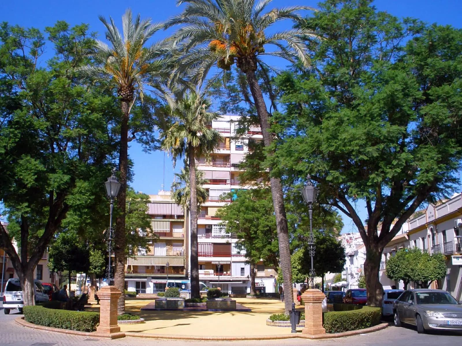 Plaza del Duque de Alcalá de Guadaíra con palmeras altas, árboles de sombra y farolas, y al fondo un bloque de pisos de seis plantas con toldos en los balcones