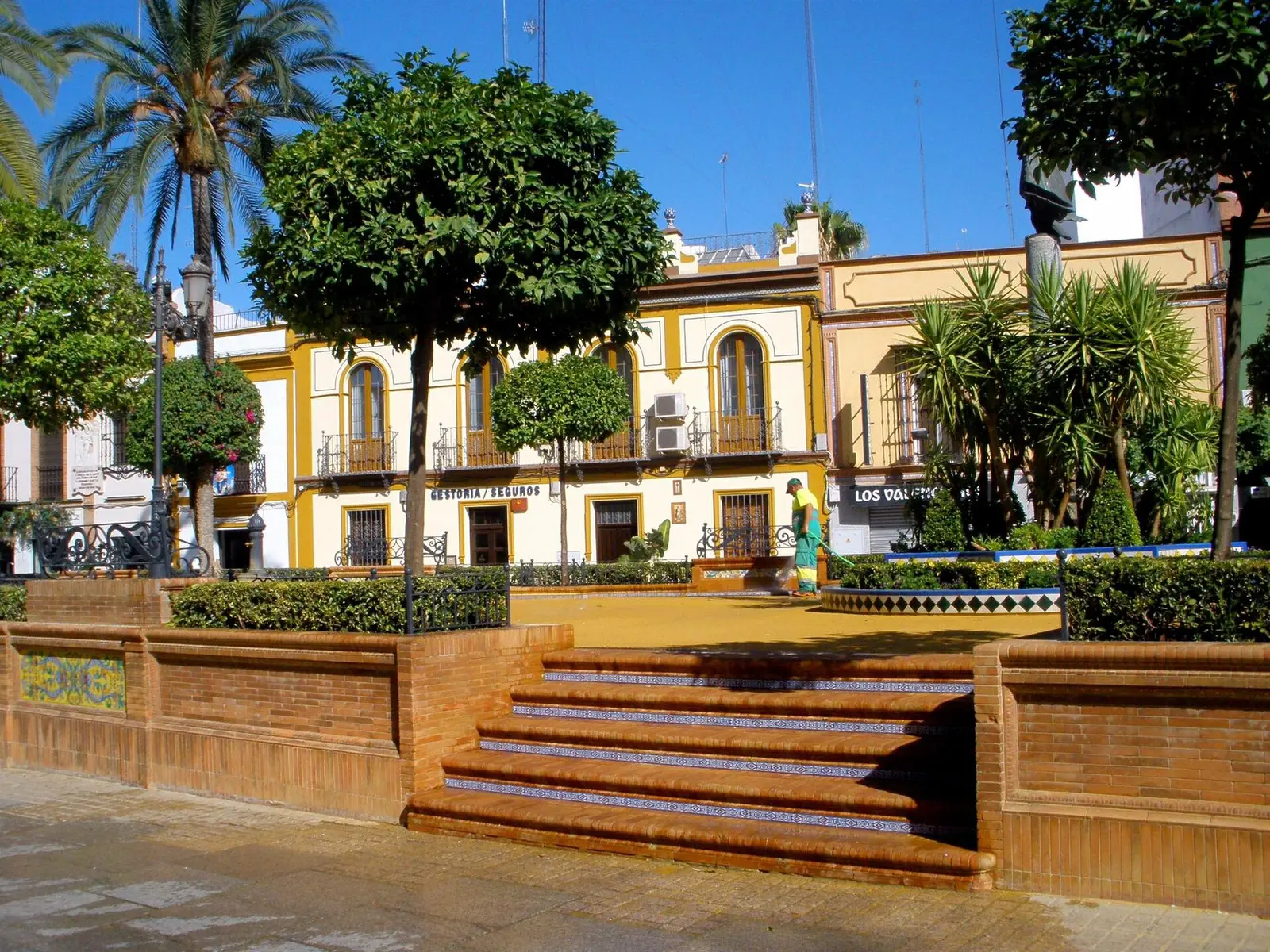 Plaza de la Plazuela de Alcalá de Guadaíra con naranjos y escalinata de ladrillo, y detrás una casa de dos plantas pintada de blanco y amarillo con balcones de forja y aparatos de aire acondicionado en la fachada