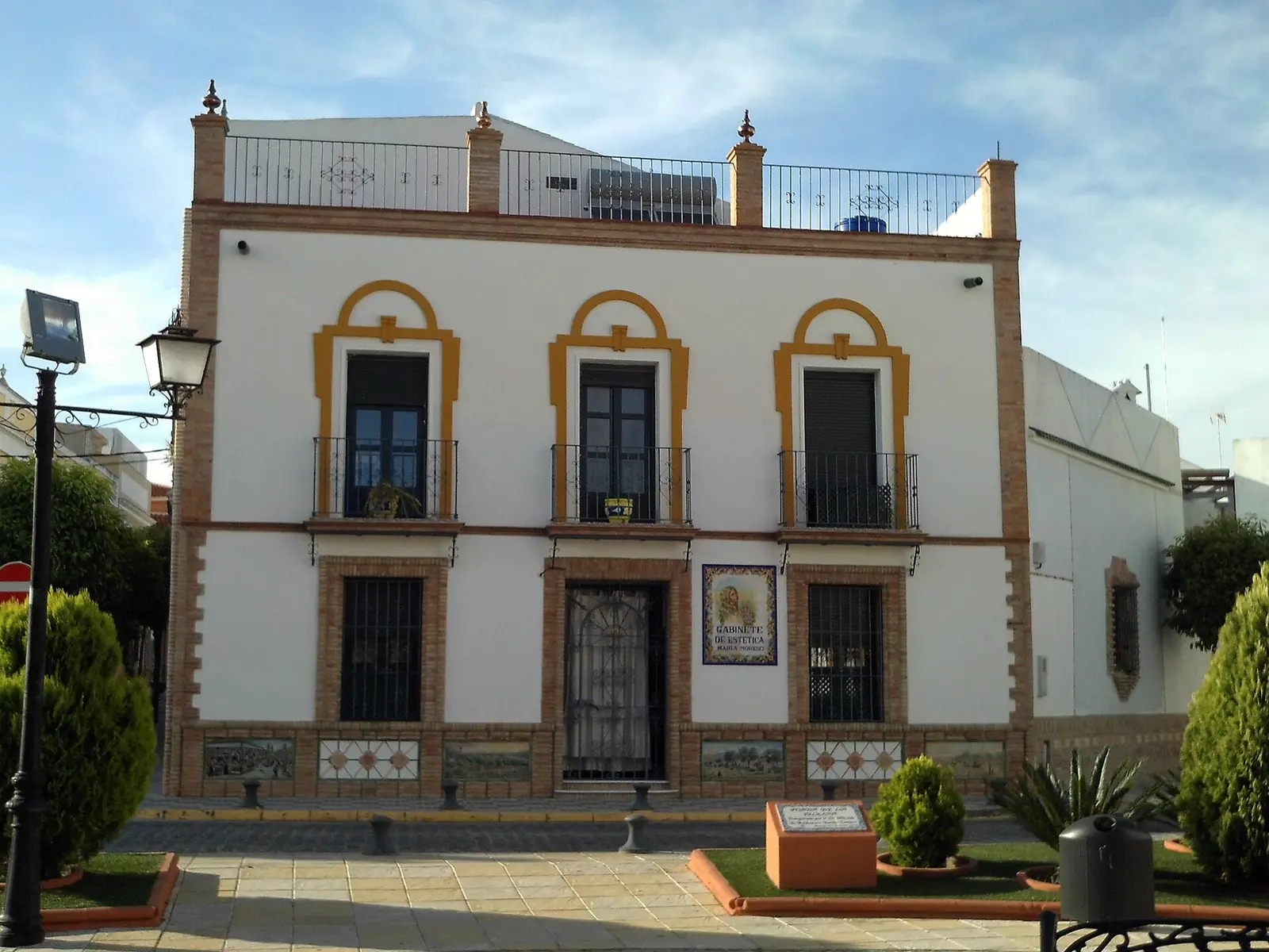 Casa tradicional de dos plantas en la plaza de la Iglesia de Bormujos, con fachada blanca, recercados de ladrillo, balcones de forja, zócalo de azulejo y una máquina de aire acondicionado en la azotea