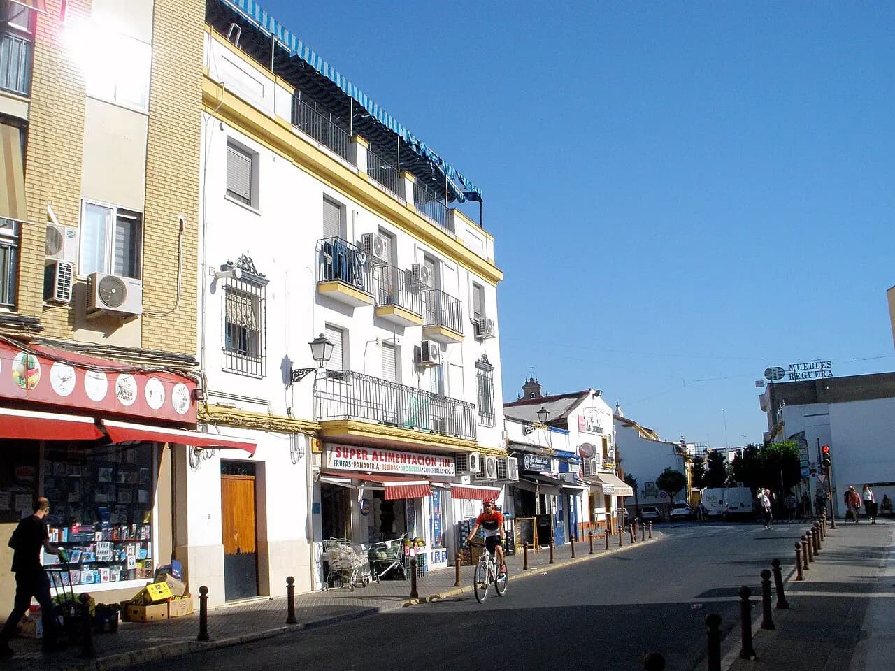 Calle comercial de Camas con un edificio blanco de tres plantas, balcones de forja y aparatos de aire acondicionado en la fachada, y un ciclista pasando