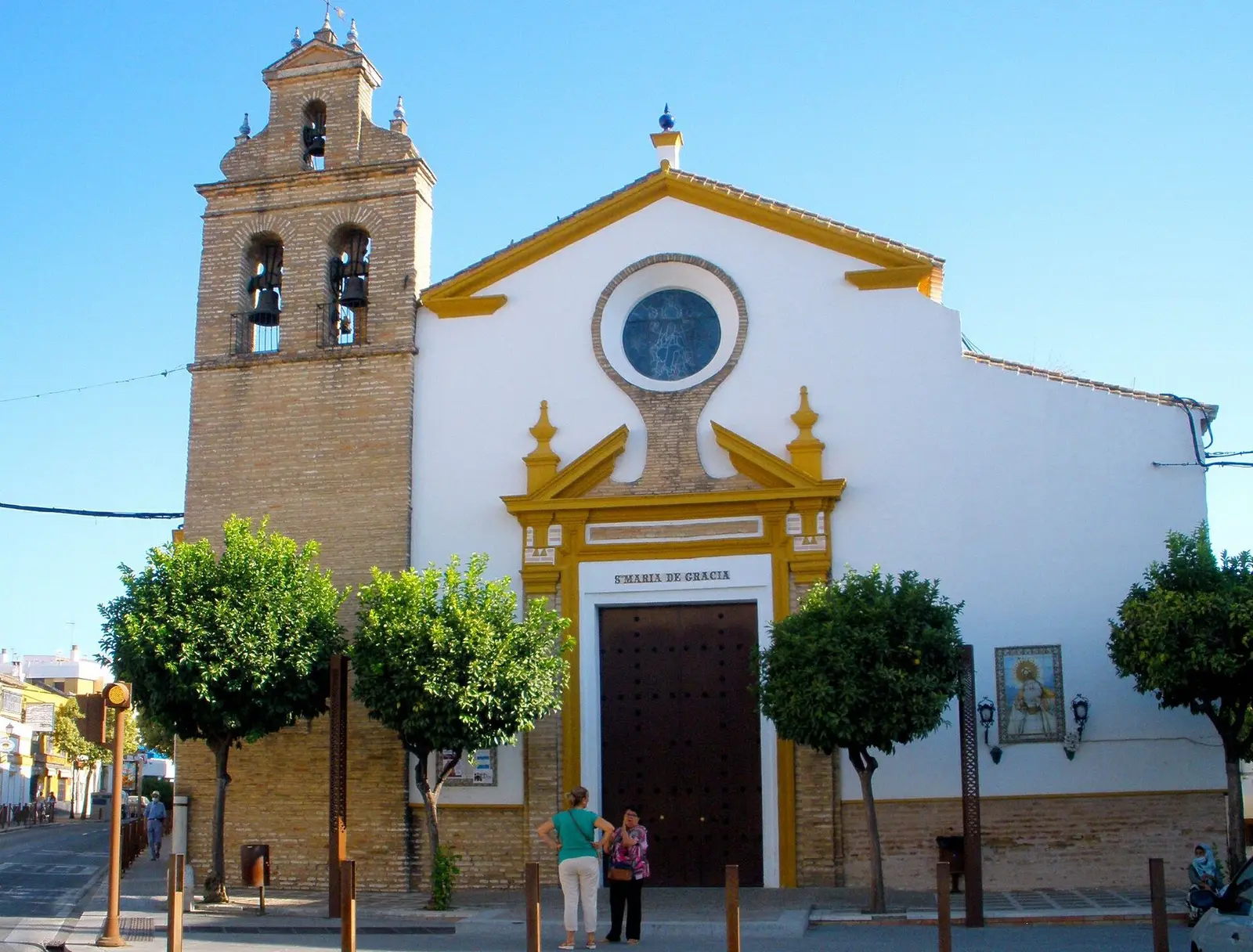 Fachada encalada de la iglesia de Santa María de Gracia de Camas, con portada de molduras amarillas, óculo sobre la puerta y torre campanario de ladrillo