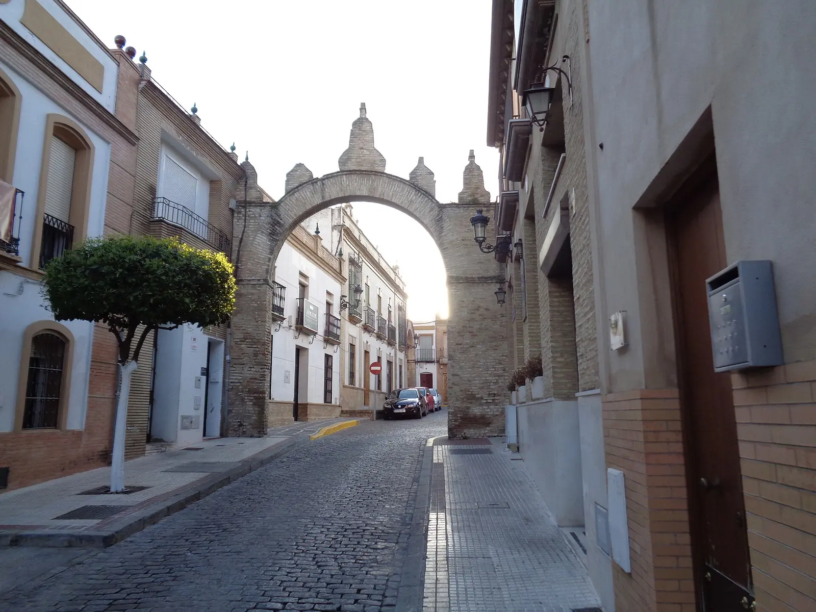 Calle adoquinada del casco de Castilleja de la Cuesta que pasa bajo un arco de ladrillo rematado con pináculos, entre casas de dos y tres plantas con balcones de forja