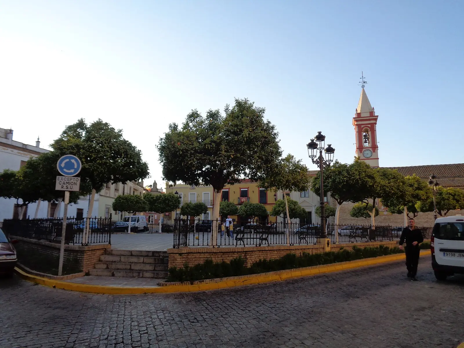 Plaza de Castilleja de la Cuesta con naranjos y farola de forja tras una verja, casas de dos plantas al fondo y el campanario rojo y blanco de una iglesia con reloj