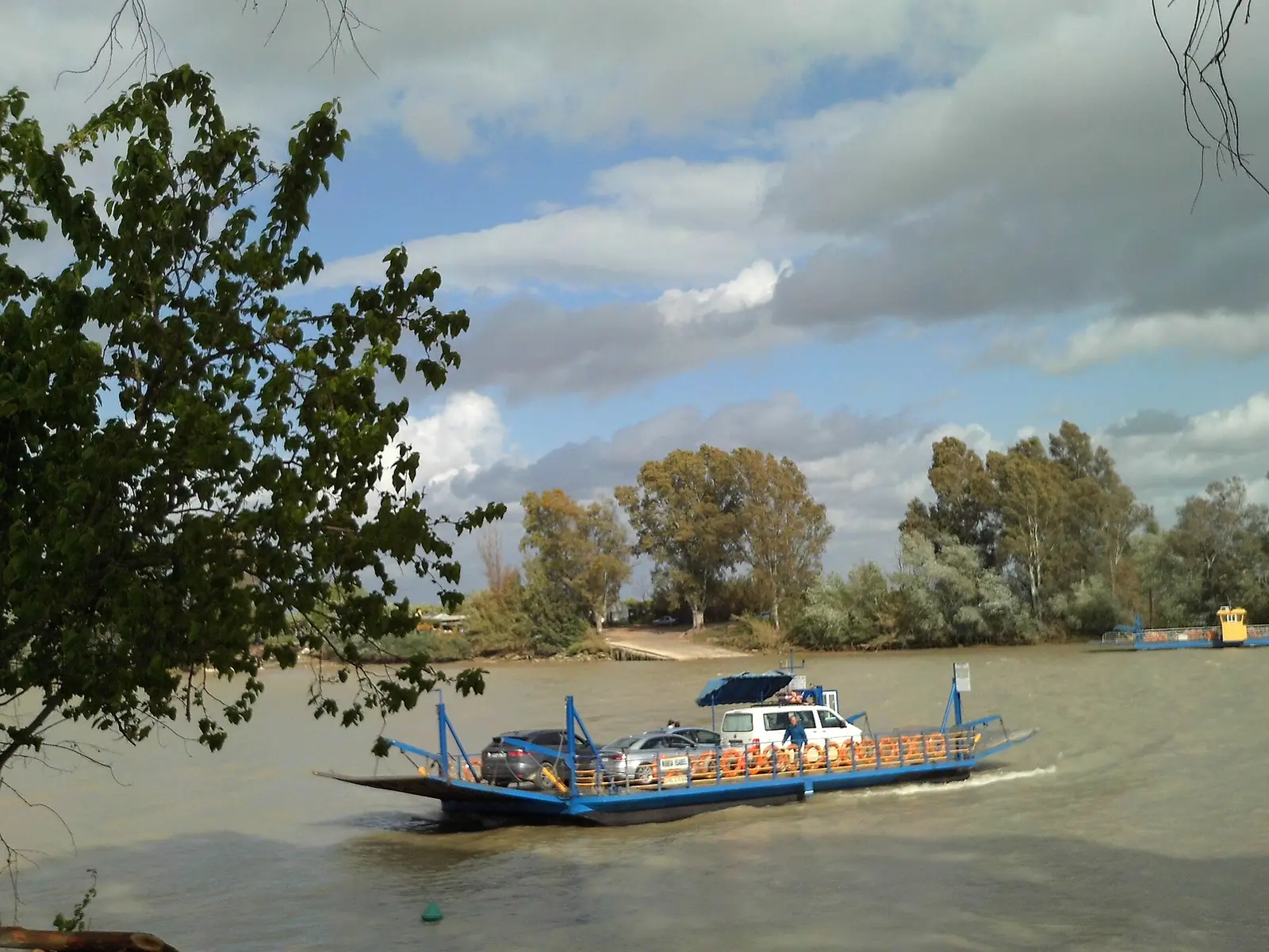 Barca de pasaje azul cruzando el Guadalquivir con varios coches a bordo, con la orilla arbolada de enfrente y un cielo con nubes