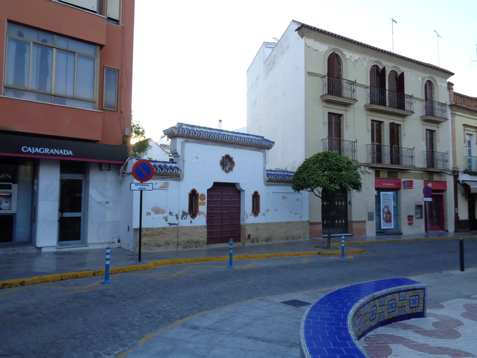 Esquina de la plaza de la Constitución de Dos Hermanas: casa baja encalada con portón de madera y remate de teja vidriada azul, junto a un edificio de tres plantas con balcones de forja y un bloque moderno de ladrillo
