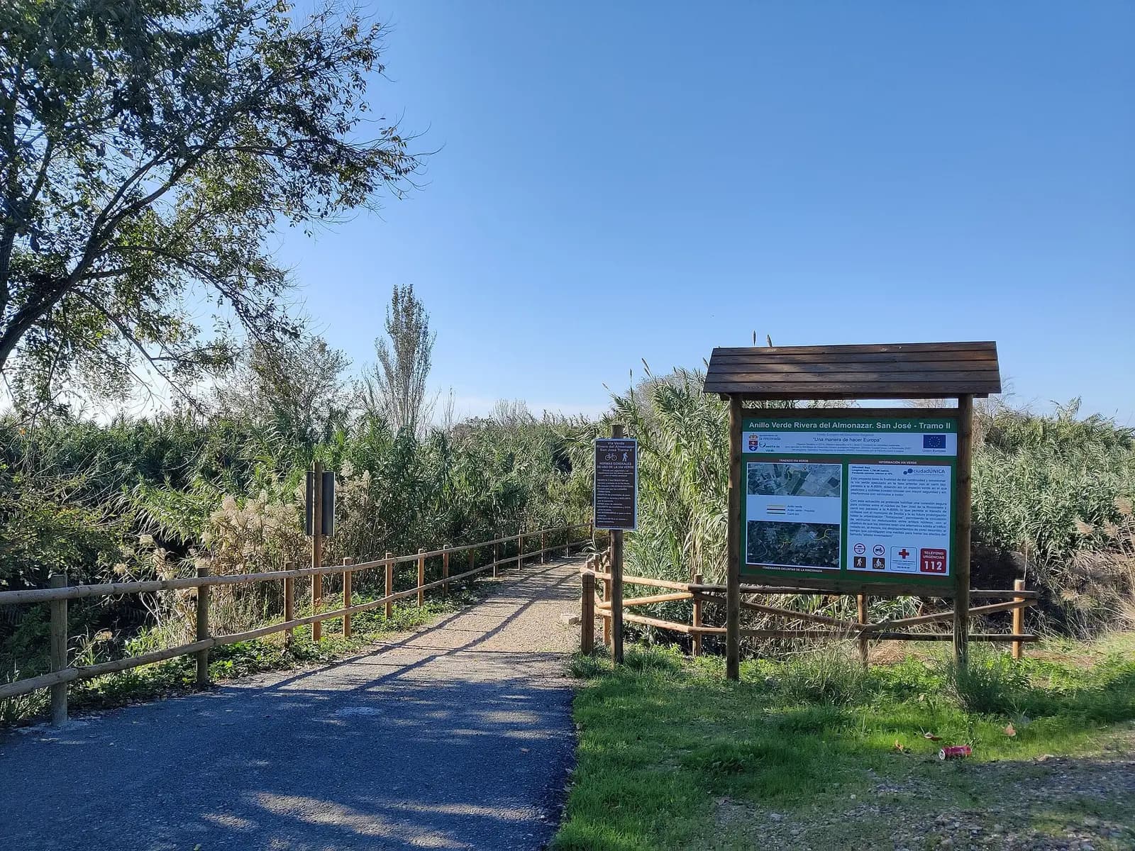 Camino de tierra con barandilla de madera junto a un cañaveral y un panel informativo del Anillo Verde Rivera del Almonazar, tramo II, en San José