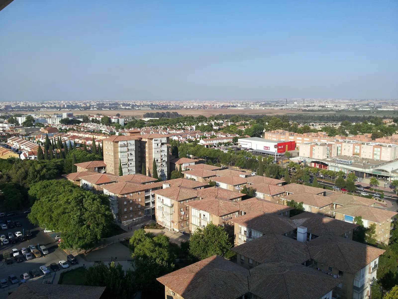 Vista aérea de Ciudad Aljarafe con bloques de ladrillo y tejados de teja a cuatro aguas, hileras de casas adosadas detrás y Sevilla al fondo