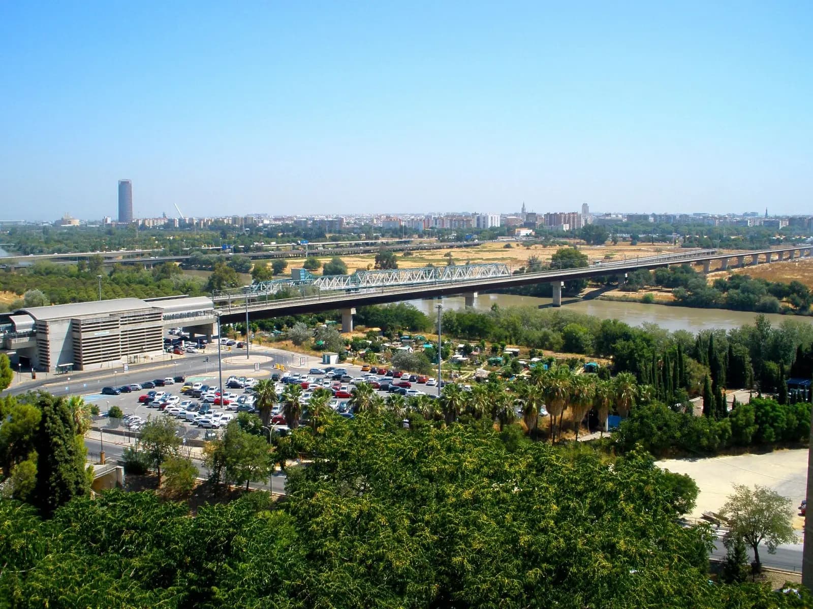 Vista desde lo alto de San Juan de Aznalfarache: un aparcamiento y un edificio de cubierta curva en primer término, puentes sobre el Guadalquivir y Sevilla al fondo con la Torre Sevilla